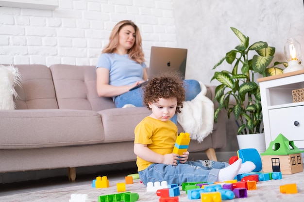 Mutter, Freiberuflerin, die Laptop verwendet, sitzt auf der Couch, Tochter der Tochter spielt mit Spielzeug auf dem Boden und arbeitet von zu Hause aus