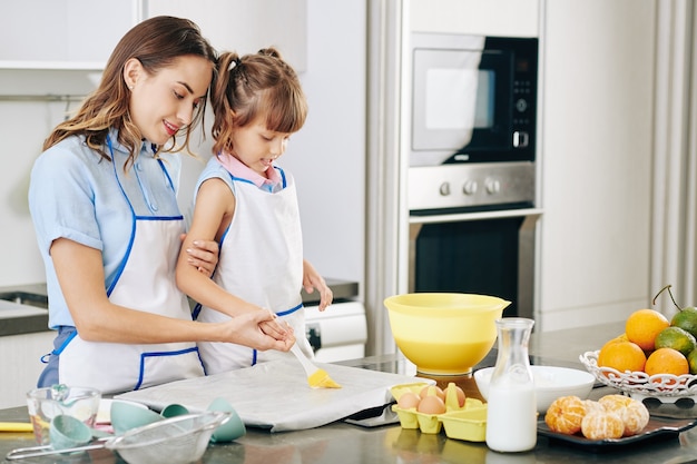 Mutter führt die Hand der kleinen Tochter, wenn sie ihr beibringt, wie man weiche Butter auf ein mit Backpapier bedecktes Tablett aufträgt