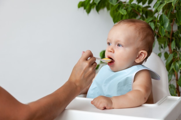 Mutter füttert entzückendes blondes Baby im blauen Lätzchen von einem Löffel