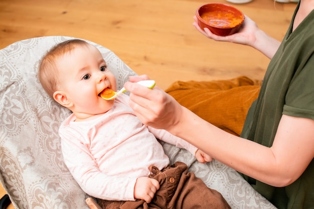 Mutter füttert lächelndes Babygemüsepüree von einem Plastiklöffel zu Hause.