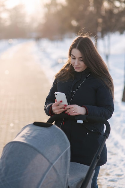 Mutter geht mit Baby im Kinderwagen Winterpark glückliche Frau mit ihrer Babymutter mit Telefon im Winter