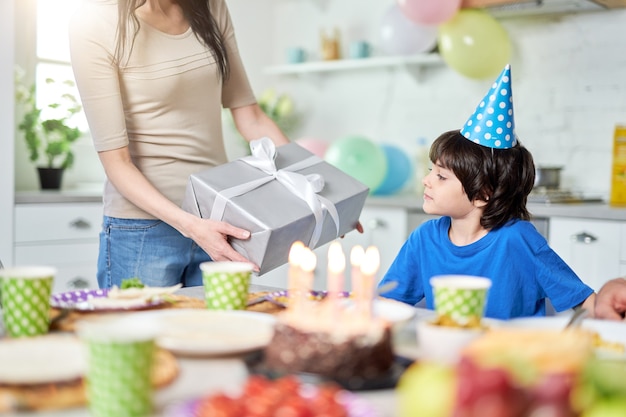 Mutter gibt ihrem schönen kleinen Jungen eine Geschenkbox, während sie zu Abend isst und den Geburtstag des Kindes zu Hause feiert. Selektiver Fokus. Familie, Feierkonzept