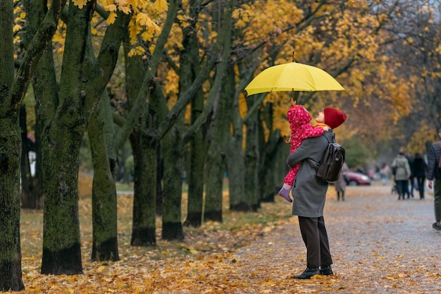 Mutter hält ein kleines Kind in ihren Armen, während sie unter einem gelben Regenschirm in einem Herbstpark vor einem Hintergrund aus gelbem Laub steht