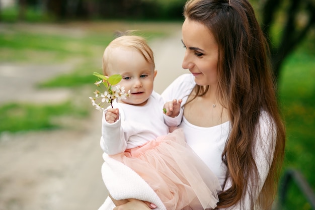 Mutter hält ihre kleine Tochter in ihren Armen zwischen blühenden Bäumen. Mama und ihr kleines Baby trugen ein rosa Kleid im Familienlook.