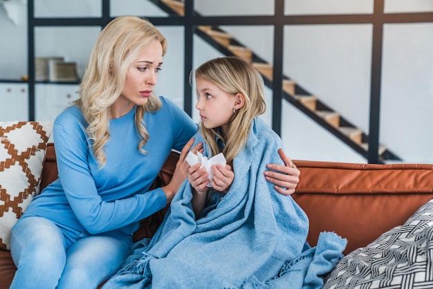 Mutter hilft ihrer kleinen Tochter mit ihrer Krankheit zu Hause auf dem Sofa