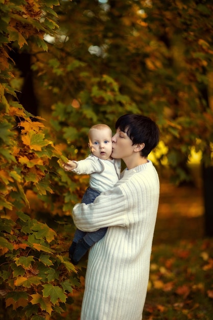 Mutter in einem leichten Pullover mit einem kleinen Kind im Arm vor dem Hintergrund des goldenen Herbstes