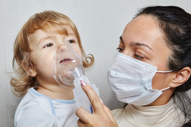 Mutter in medizinischer Maske hilft ihrer kleinen Tochter beim Atmen mit Hilfe eines Verneblers, der Atemwegserkrankungen behandelt.