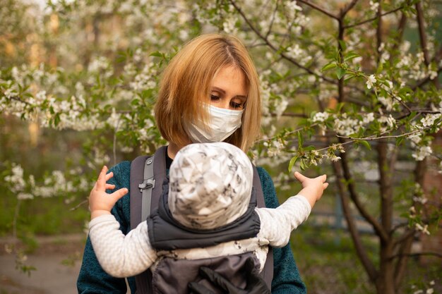 Mutter in medizinischer maske mit kleinem sohn im park spazieren in der nähe von weiß blühendem baum covid-konzept