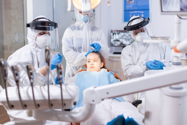 Mutter in PPE-Anzug gekleidet, die Tochter während der zahnärztlichen Behandlung tröstet. Stomatologie-Team, das während der Covid19-Prozedur an Kinderzähnen einen PPE-Anzug trägt.