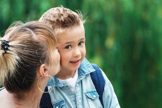 Mutter küsst Sohn in Schach, bevor sie zur Schule geht. stilvoller kleiner Schuljunge in einem blauen Hemd mit einem Rucksack im Park