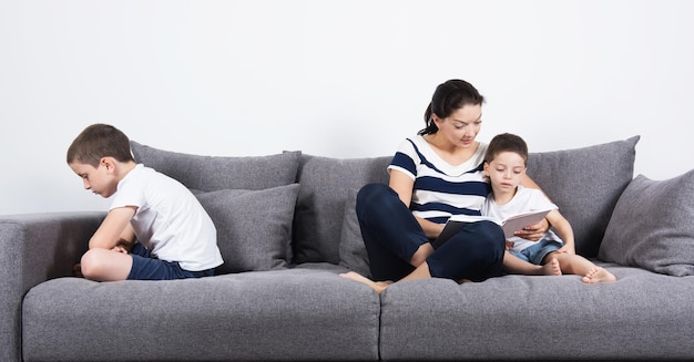 Mutter liest mit ihrem Sohn ein Buch und ein anderer Sohn ist auf der gegenüberliegenden Seite des Sofas beleidigt