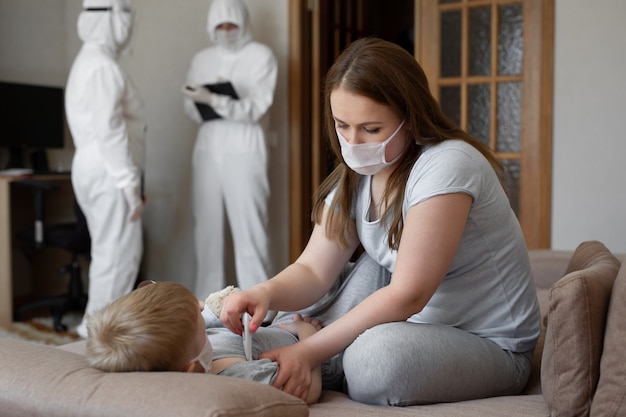 Mutter misst die Temperatur des Babys. Ärzte in Schutzanzügen bei kranken Patienten zu Hause. Coronavirus (COVID-19