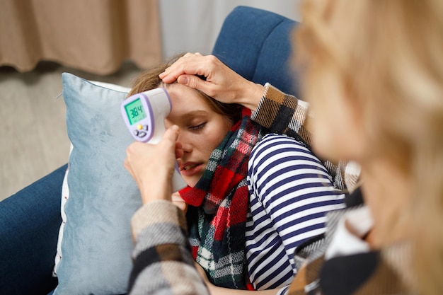 Mutter misst die Temperatur ihrer an Grippe erkrankten Tochter, die zu Hause auf dem Sofa liegt