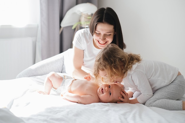 Mutter mit dem lockigen Mädchen, das Babybruder auf dem Bett küsst