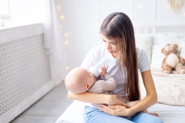 Mutter mit einem Baby im Arm füttert Milch aus einer Flasche auf dem Bett zu Hause in einem hellen Raum das Konzept der Babynahrung Mutter macht sich Sorgen um ein kleines Kind