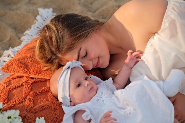 Mutter mit einem neugeborenen Baby liegt am Strand, weiße Kleider, Blumen liegen in der Nähe