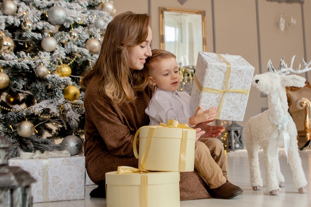 Mutter mit ihrem kleinen Sohn, der ein Weihnachtsgeschenk im gemütlichen Wohnzimmer öffnet. Familienurlaub