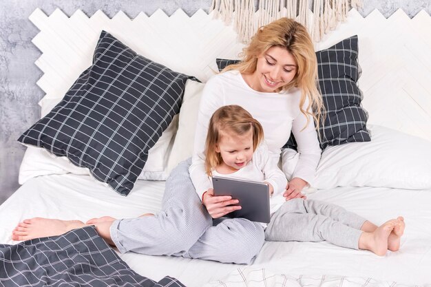 mutter mit ihrer kleinen tochter liegt im bett und schaut auf das tablet