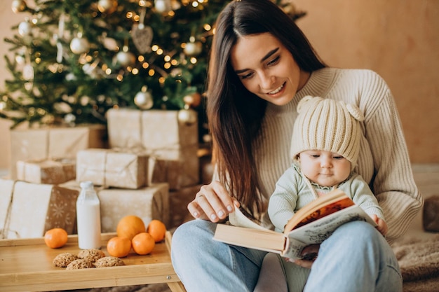 Mutter mit ihrer kleinen Tochter liest ein Buch am Weihnachtsbaum