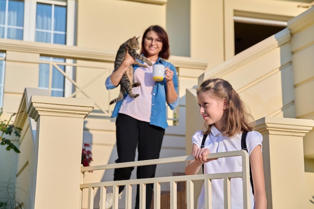 Mutter mit Katze im Arm auf Außentreppe morgens zu Hause mit jugendlicher Tochter, die zur Schule geht