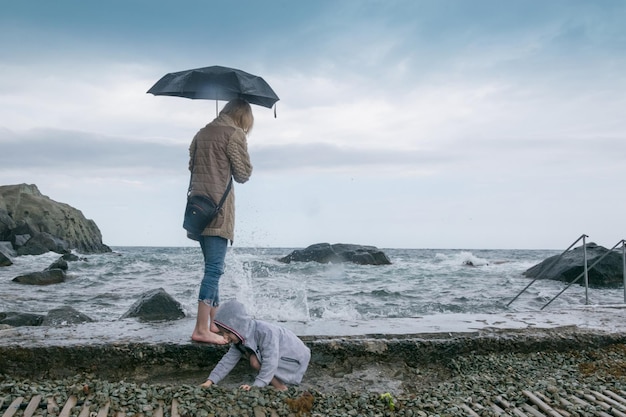 Mutter mit Kind kleiner Junge an einem steinigen Strand bei schlechtem Wetter am Strand das Meer Spaziergang entlang der Küste