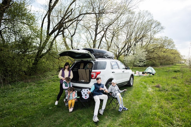 Mutter mit Kindern sitzt beim Picknick auf einem Stuhl vor dem Auto