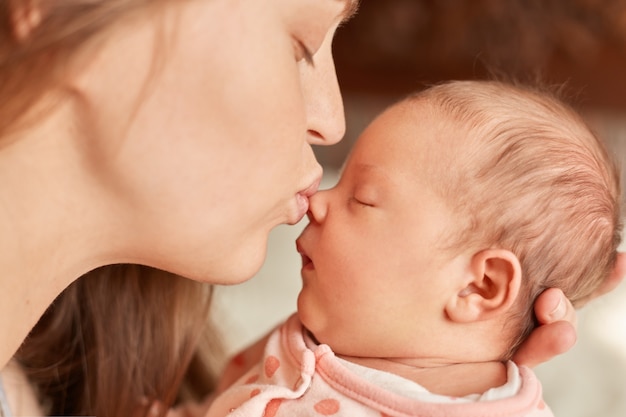 Mutter mit Neugeborenem posiert drinnen, Mutter küsst ihr schläfriges Kind, weibliche geschlossene Augen und Schmollmund, genießt es, Zeit mit ihrem Kind zu verbringen, Mutterschaft.