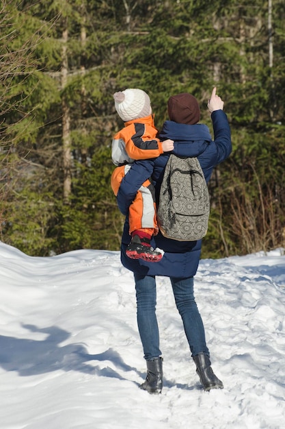 Mutter mit Sohn in Armen und Rucksack steht vor dem Hintergrund von Nadelwald und schneebedeckter Straße Wintertag