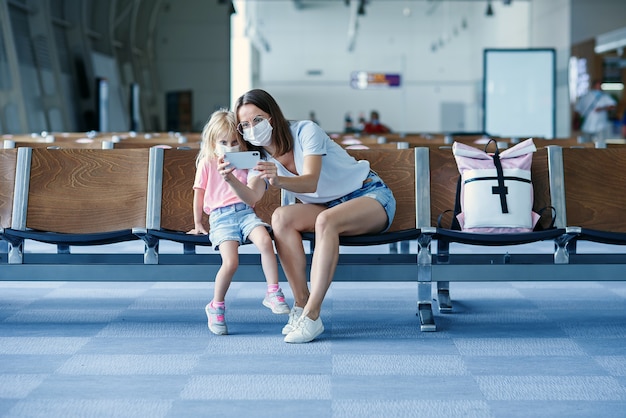 Mutter mit Tochter in Masken wartet auf ihren Flug am Flughafen Frau mit kleinem Mädchen in