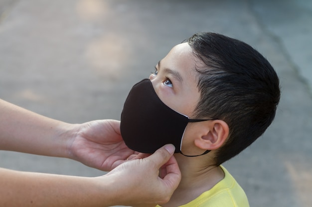 Mutter trägt eine dunkle Gesichtsschutzmaske für ihren Sohn. Schwarzhaariger asiatischer Junge trägt mit seiner Mutter eine schwarze Gesichtsmaske. Hintergrund für das Covid-19-Gesundheitspflege- oder PM 2.5-Mikrostaub-Verschmutzungsproblemkonzept.