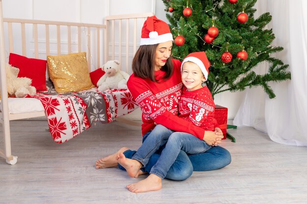 Mutter umarmt ein Kind in roten Pullovern neben dem Weihnachtsbaum