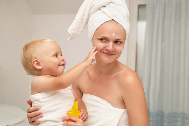 Mutter und Baby in weißen Handtüchern tragen nach dem Baden Sonnencreme oder After Sun Lotion auf