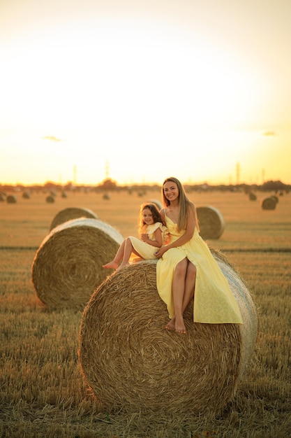 Mutter und blonde Tochter sitzen bei Sonnenuntergang auf einem Heuhaufen auf einem Feld Mutter und Kind haben Spaß auf dem Bauernhof