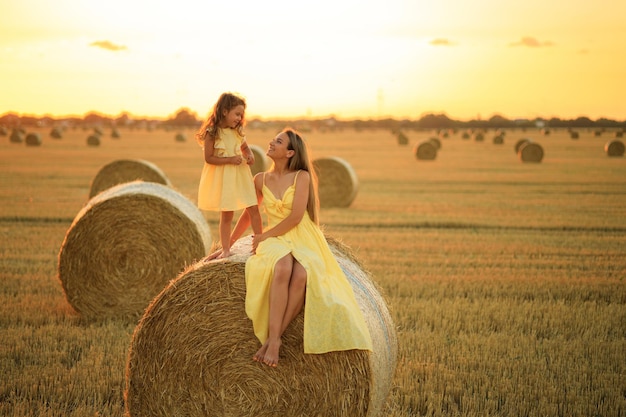 Mutter und blonde Tochter sitzen bei Sonnenuntergang auf einem Heuhaufen auf einem Feld Mutter und Kind haben Spaß auf dem Bauernhof