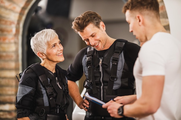 Mutter und ihr erwachsener Sohn sprechen nach dem EMS-Training im Fitnessstudio mit dem Personal Trainer.