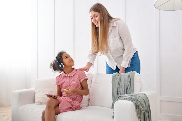 Mutter und ihre kleine Tochter in Kopfhörern auf dem Sofa im Wohnzimmer. Mutter und weibliches Kind Freizeit in ihrem Haus zusammen, gute Beziehung, elterliche Fürsorge