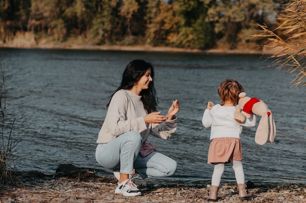 Mutter und Kind am Strand auf dem Hintergrund eines blauen Sees, Mutter spielt mit ihrer Tochter auf dem See. Herbst Natur