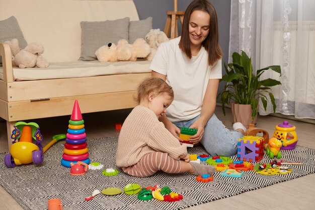 Mutter und Kind bauen aus bunten Plastikblöcken lächelnde Frau und ihre Tochter im Spielzimmer, die Ziegelkonstruktionen für die frühe Entwicklung der Vorschulerziehung herstellen