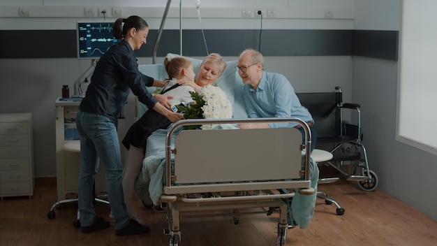 Mutter und Kind besuchen kranke Oma im Bett der Krankenstation und geben Blumenstrauß. Kranker Patient, der Besuch von Nichte und Tochter genießt und kleines Mädchen umarmt. Ältere Frau mit Familie.