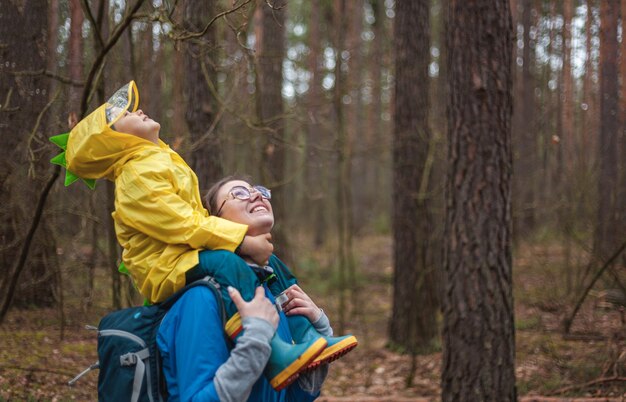Mutter und Kind gehen nach dem Regen in Regenmänteln zusammen im Wald spazieren, das Kind sitzt auf den Schultern der Mutter und schaut glücklich in den Himmel