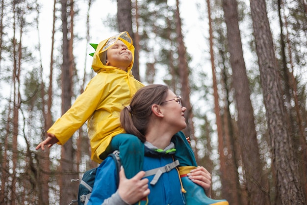 Mutter und Kind gehen nach dem Regen in Regenmänteln zusammen im Wald spazieren, das Kind sitzt auf den Schultern der Mutter und schaut glücklich in den Himmel