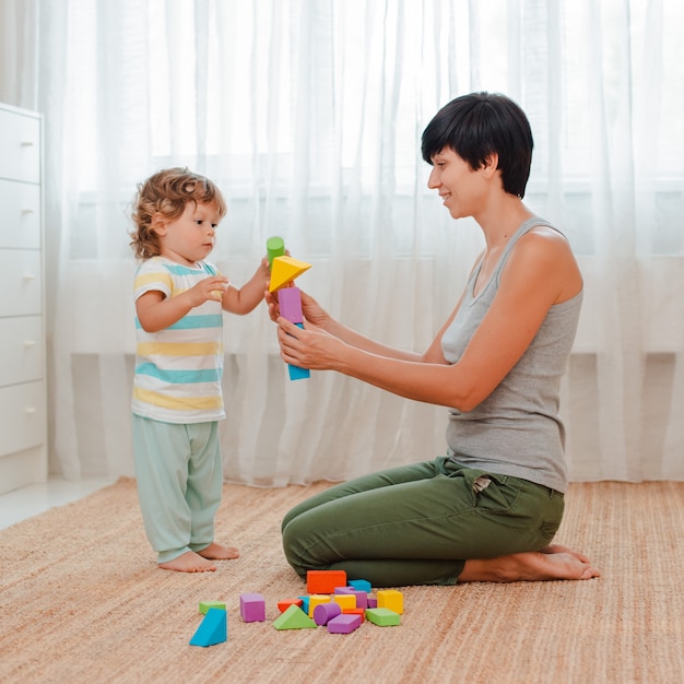 Mutter und Kind spielen im Kinderzimmer auf dem Boden. Mama und ein kleiner Junge bauen einen Turm aus farbigen Blöcken.