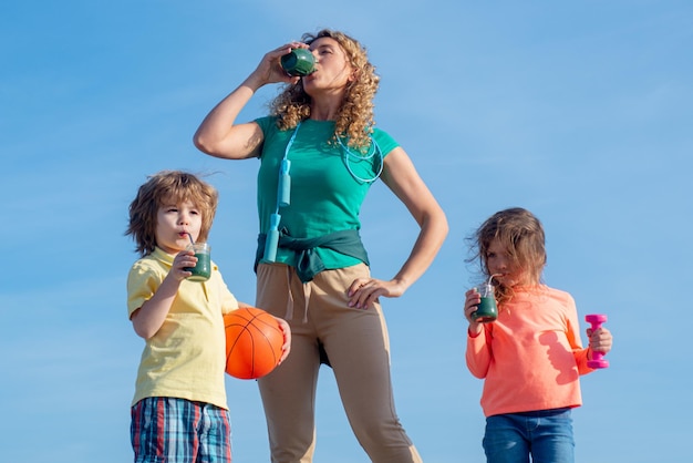 Mutter und Kinder trinken Smoothie oder Saft Familie nach Sportübungen auf blauem Himmelshintergrund kopieren Raum Fröhliche lachende Kinder und ihre schöne junge Mutter sportlich liebende Familie