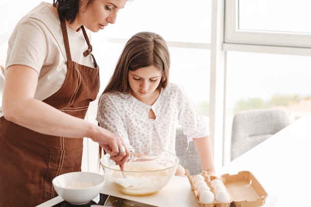 Mutter und kleine Tochter der kaukasischen Familie backen Gebäck in der Küche zu Hause und kneten Teig mit Löffel