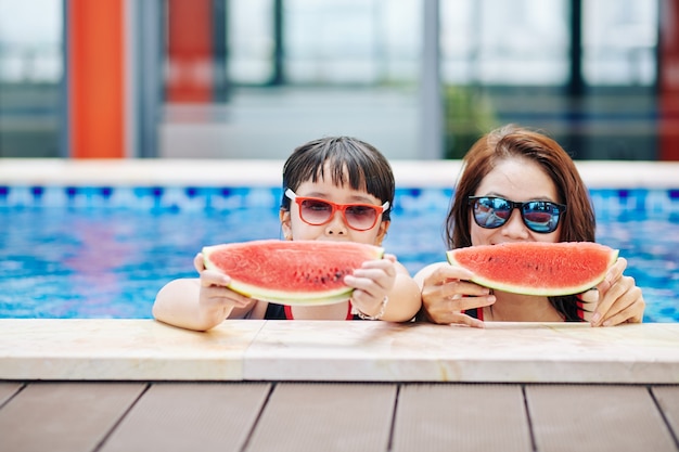Mutter und kleine Tochter mit Sonnenbrille stehen im Schwimmbad und essen süße leckere Wassermelonenscheiben