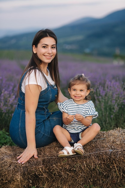 Mutter und kleine Tochter sitzen auf dem Heu auf dem Bauernhof Hintergrund des sommerlichen Lavendelfeldes Familien-Denim-Stil