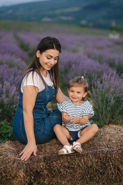 Mutter und kleine Tochter sitzen auf dem Heu auf dem Bauernhof Hintergrund des sommerlichen Lavendelfeldes Familien-Denim-Stil