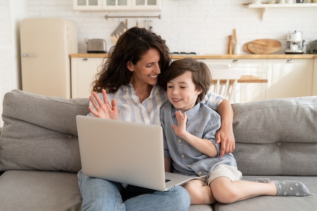 Mutter und kleines Kind, die während des Videoanrufs auf dem Laptop grüßen, winken der Computer-Webcam mit den Händen, die zu Papa lächeln