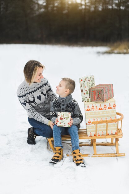 Mutter und Sohn draußen sitzen auf Holzschlitten mit Weihnachtsgeschenken verziert