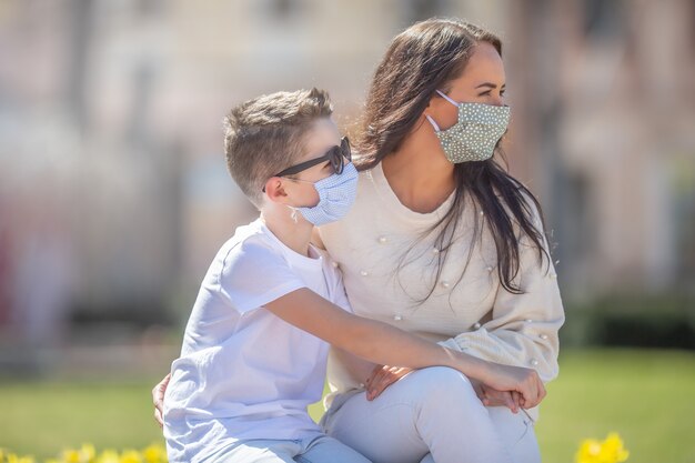 Mutter und Sohn mit Gesichtsmasken im Gesicht schauen an einem sonnigen Tag im Park beide nach rechts.